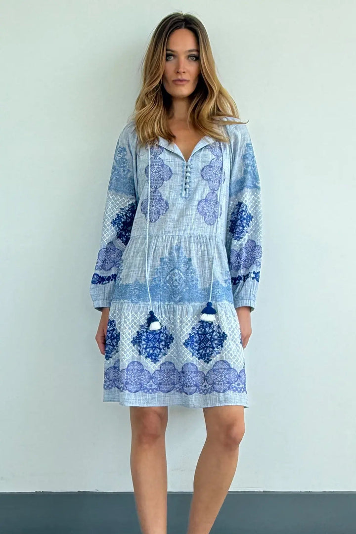 A woman modeling a light blue and blue patterned long-sleeve mini dress with a gathered waist and front button placket, standing against a plain light wall on a wood-look floor.