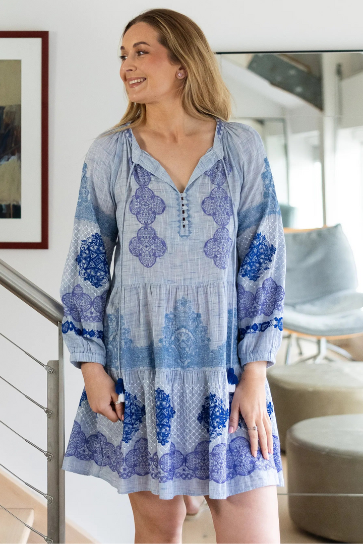 A woman wearing a light blue and purple patterned tunic dress stands in a modern home interior next to a staircase railing.