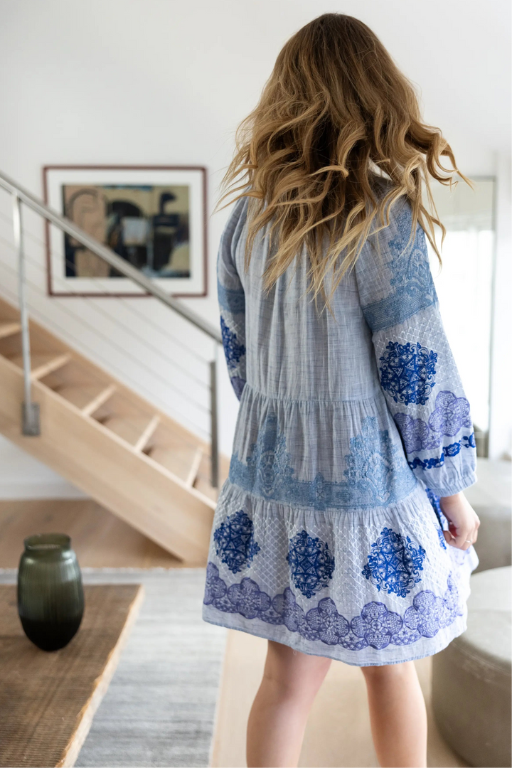 A woman viewed from behind wearing a light blue and white tiered gauze dress with blue floral lace appliques, standing in a modern home interior with a staircase in the background.