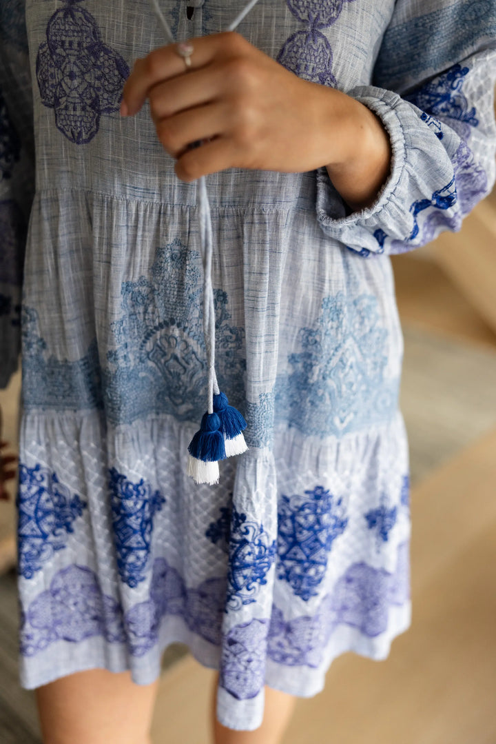A close-up of a person wearing a tiered bohemian-style dress with a blue and white batik print, holding a drawstring with tassel details.