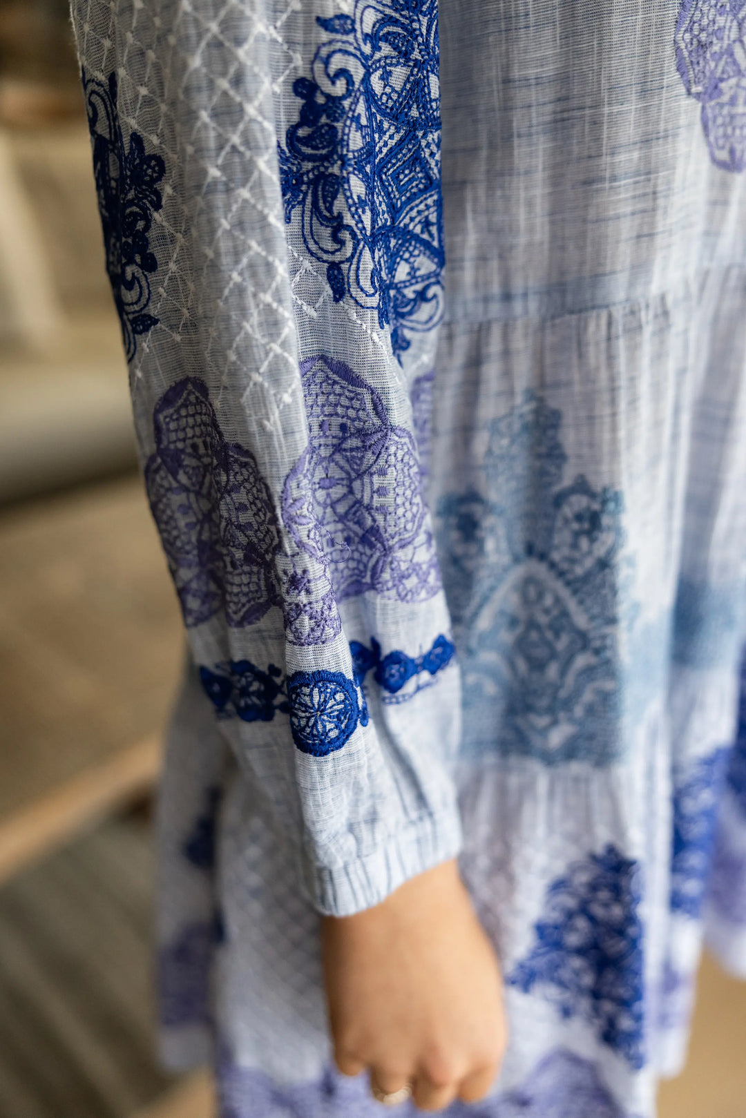 A close-up of a light blue and white printed dress with blue floral and geometric patterns, featuring a long sleeve and a visible hand.