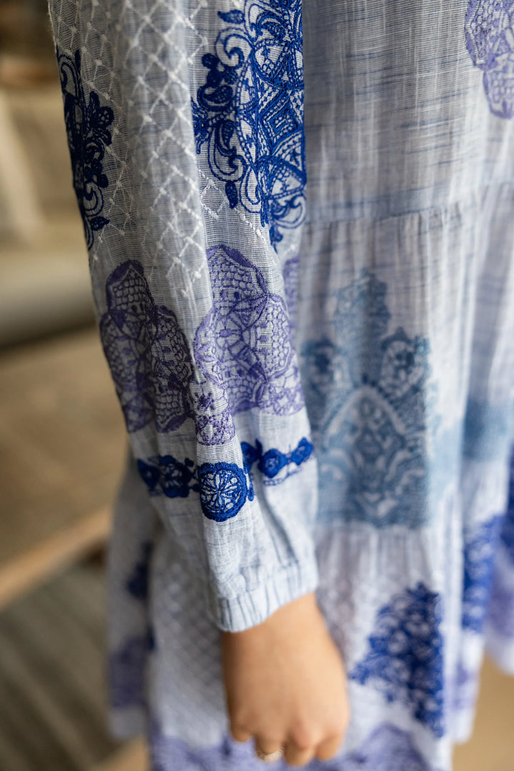 A close-up of a light blue and white printed dress with blue floral and geometric patterns, featuring a long sleeve and a visible hand.