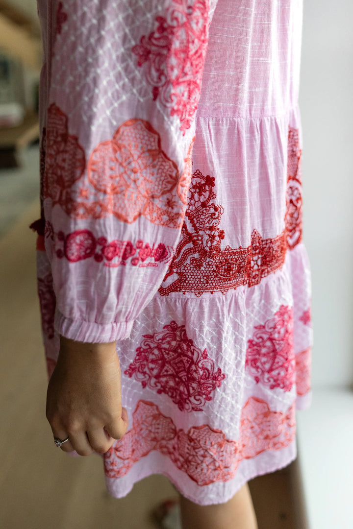A close-up of a person wearing a tiered pink dress with red and orange floral embroidery and lace-like printed patterns, shown from the side with one hand visible.