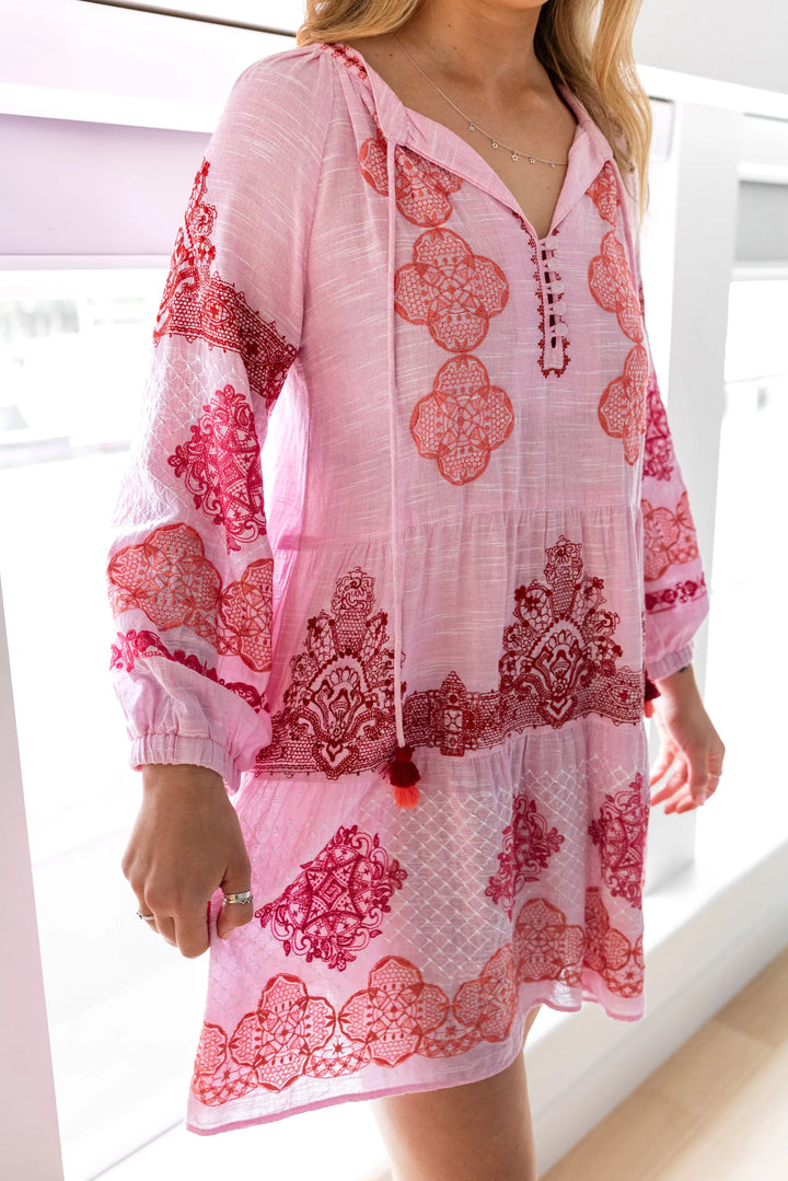A woman wearing a pink and red floral print kaftan dress stands on a light wood staircase in a modern home interior.