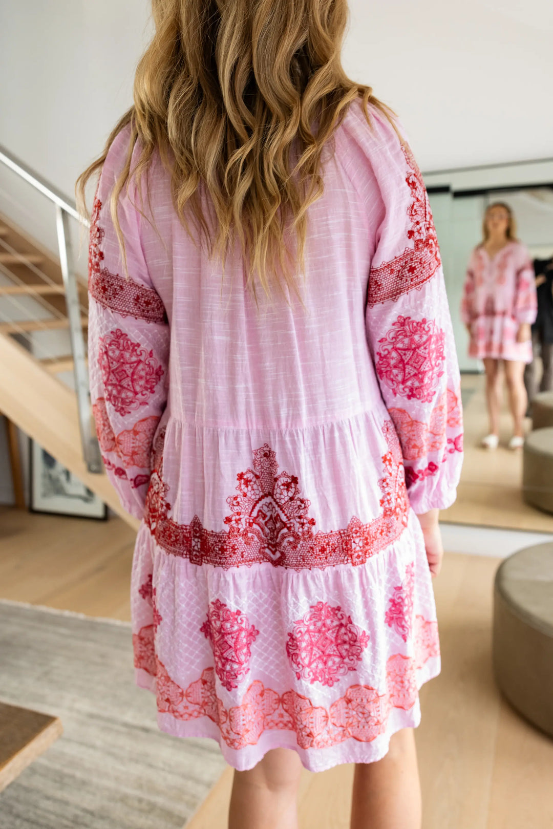 A woman wearing a light pink tiered cotton gauze dress with red and pink floral embroidery, shown from behind in a bright modern room.