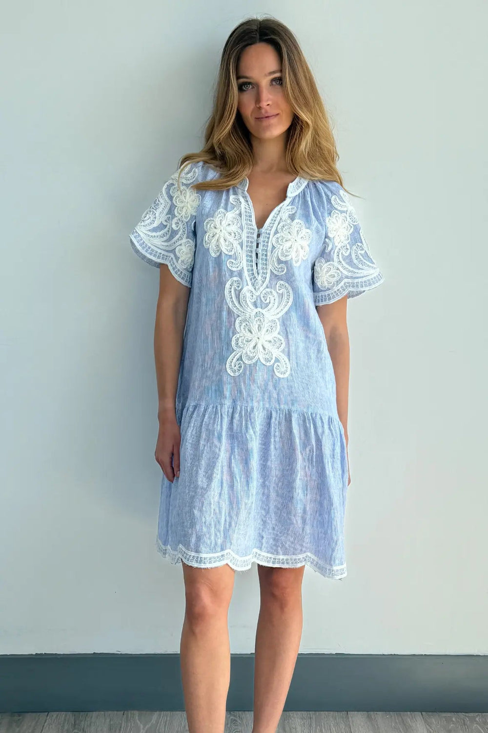 A woman modeling a light blue short-sleeve dress with white floral embroidery, standing against a light gray wall on a gray wood floor.