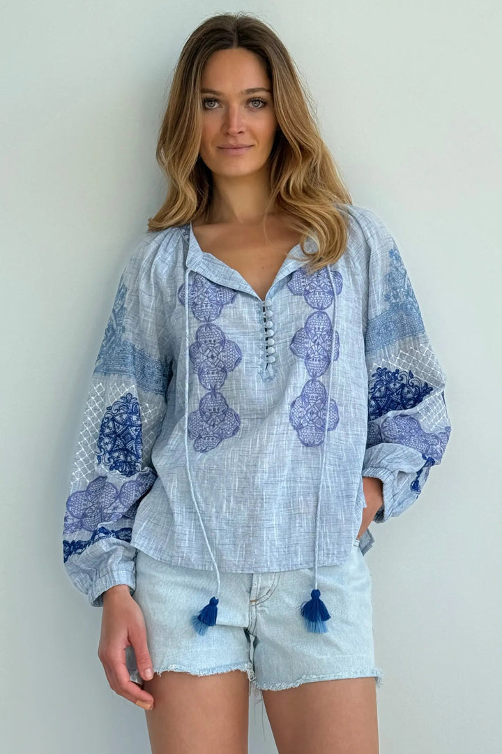 A woman modeling a light blue and blue patterned bohemian-style blouse with tassel ties, paired with light denim shorts, against a plain off-white wall.