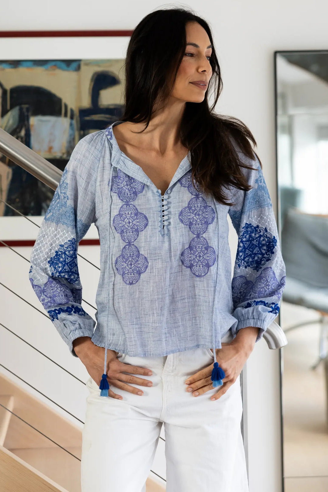 A woman stands indoors near a staircase and mirror, wearing a light blue and white patterned blouse with white pants.
