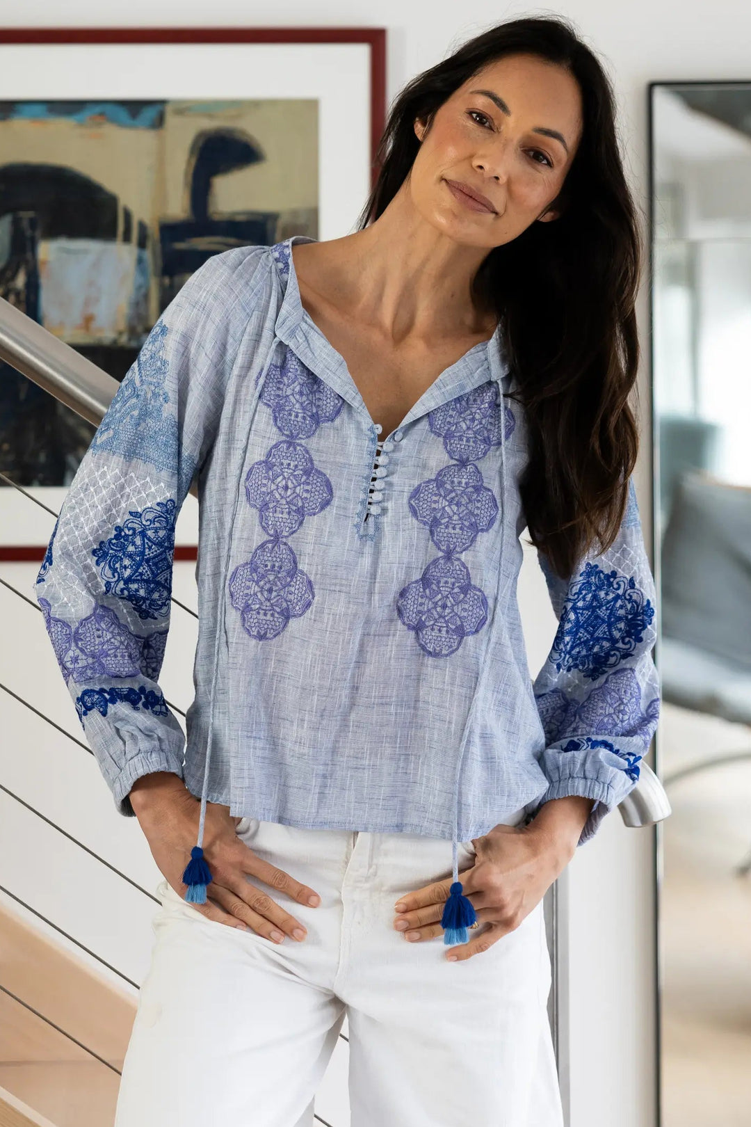 A woman wearing a light blue and purple patterned blouse with white pants, standing in a modern home interior with a staircase and mirror in the background.