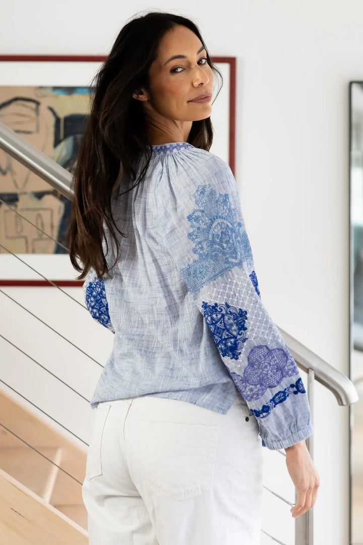 A woman wearing a light blue and white patterned blouse with long sleeves and white pants, standing on a wooden staircase in a modern home interior.