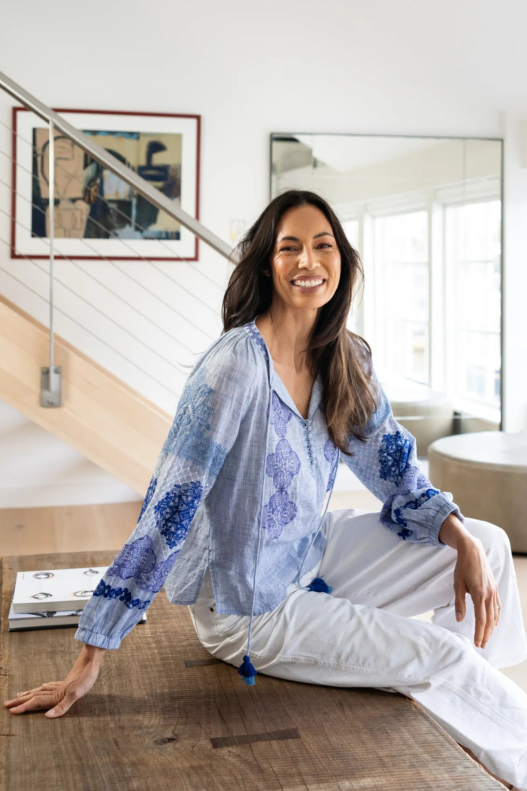 A woman with long dark hair sits smiling on a wooden coffee table in a bright, modern living room. She is wearing a light blue and white patterned long-sleeve blouse and white pants.