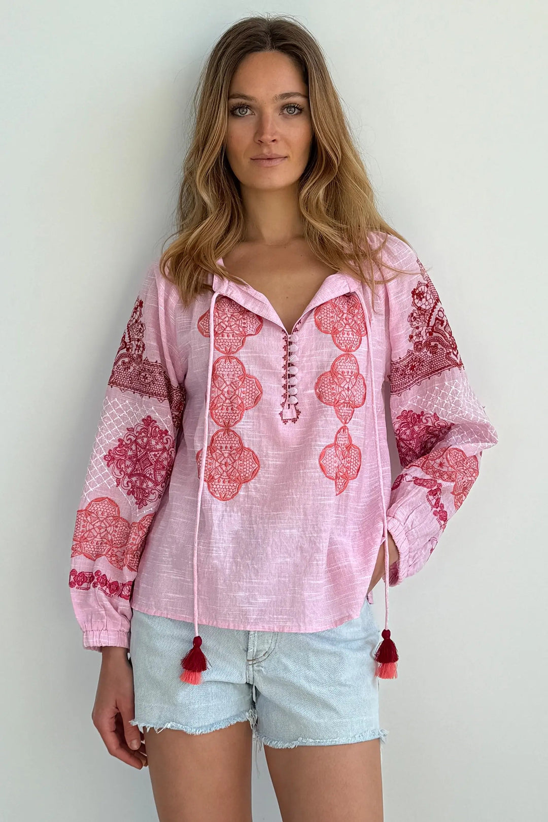 A woman modeling a light pink long-sleeve bohemian-style blouse with red and pink floral embroidery, lace-up front, and drawstring tassels, paired with light denim shorts against a plain white wall.
