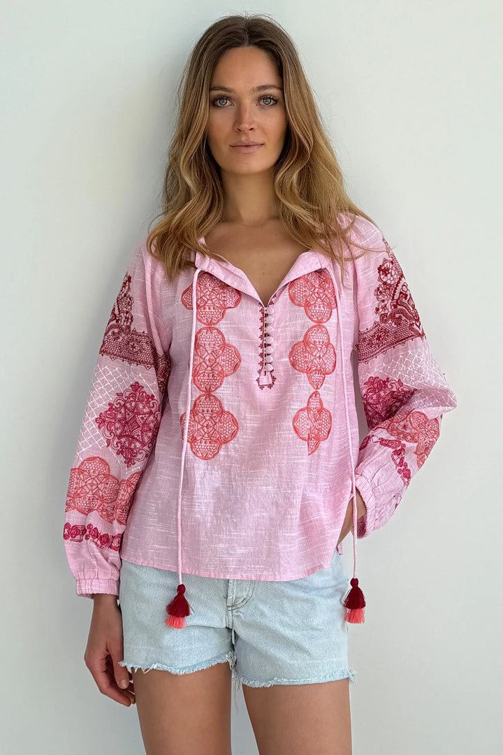 A woman modeling a light pink long-sleeve bohemian-style blouse with red and pink floral embroidery, lace-up front, and drawstring tassels, paired with light denim shorts against a plain white wall.