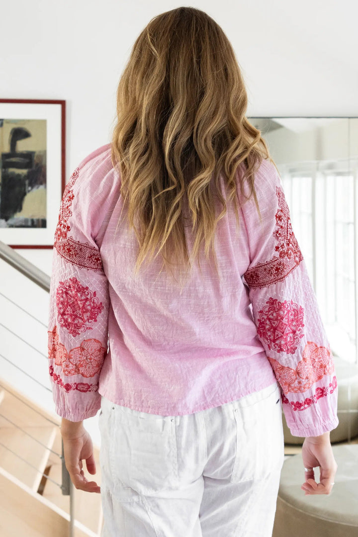 A woman viewed from behind wearing a light pink long-sleeve blouse with red floral embroidery on the sleeves and white pants, standing in a bright modern interior with a staircase and mirror in the background.