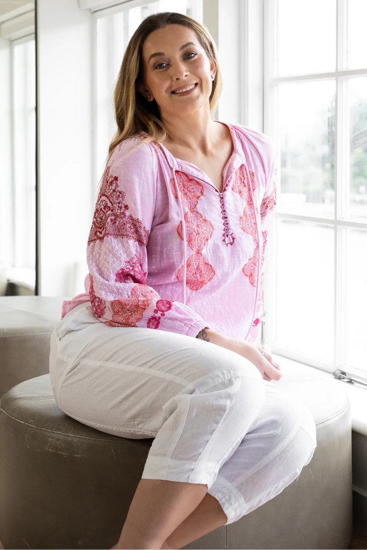 A woman wearing a pink and white patterned long-sleeve top and white cropped pants sits on a round gray ottoman in a bright, modern room with large windows and a mirror reflecting her.