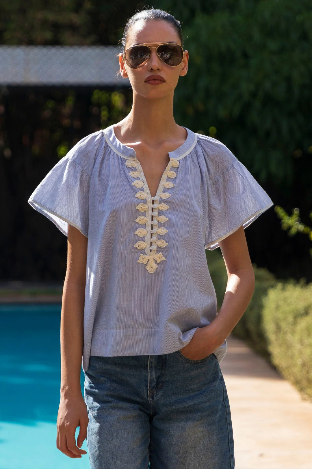 Woman wearing a light blue embroidered top and sunglasses by a poolside.