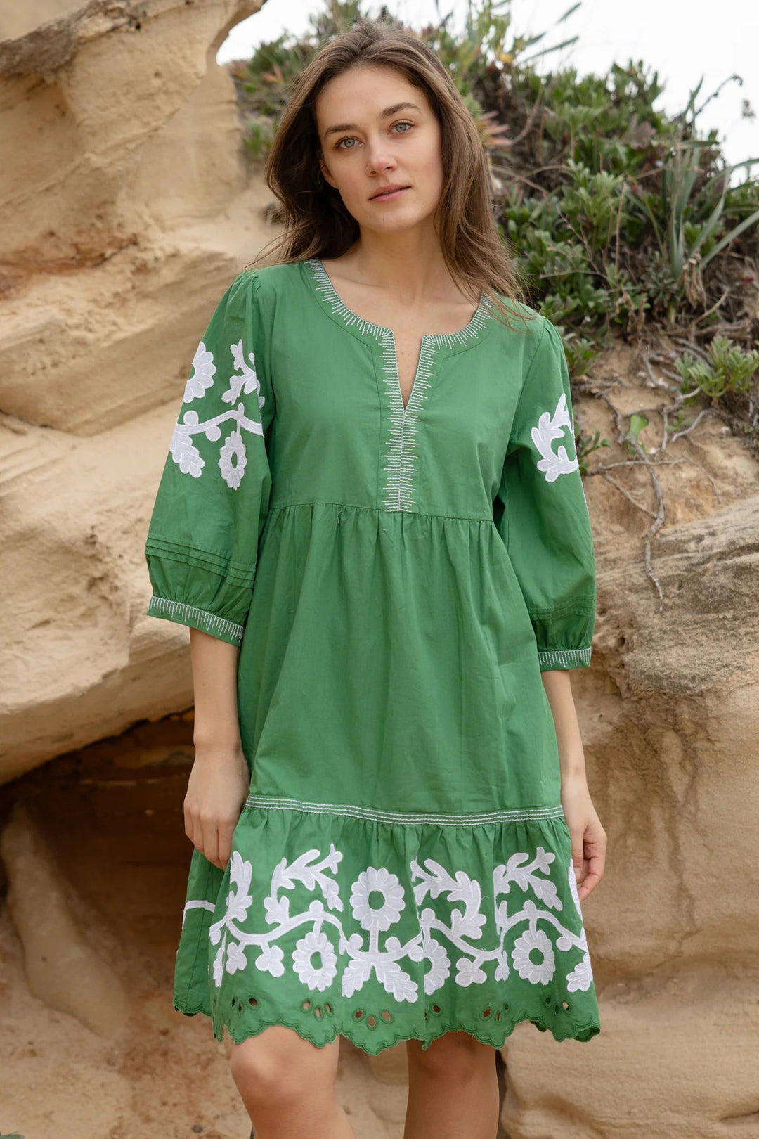 A woman wearing a green embroidered dress stands outdoors in front of sandy rock formations and greenery. The dress has white floral embroidery on the sleeves and hem, with a split neckline and elbow-length sleeves.