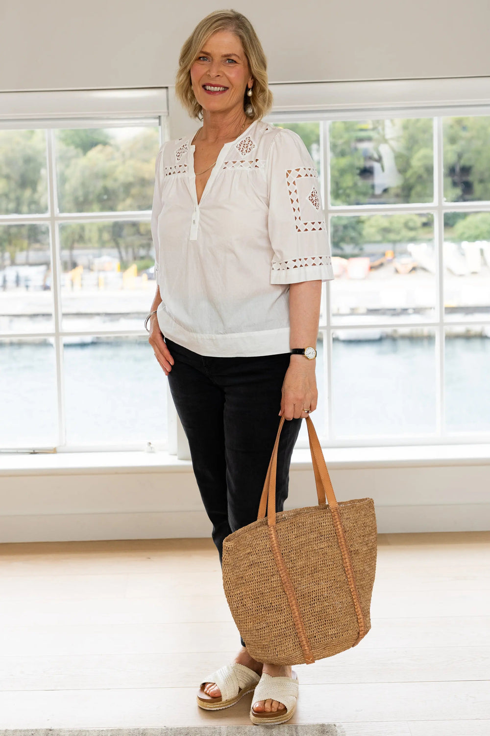 A woman stands indoors near a large window, wearing a white eyelet blouse, black pants, and white slide sandals, while holding a large woven straw tote bag with tan handles.