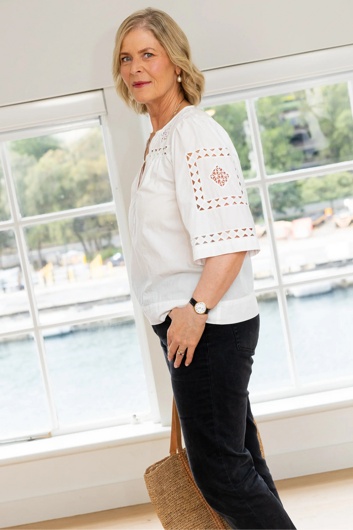 A woman wearing a white short-sleeve blouse with crochet embroidery on the sleeve and a split neckline, paired with black pants, standing in a bright room with large windows overlooking water.