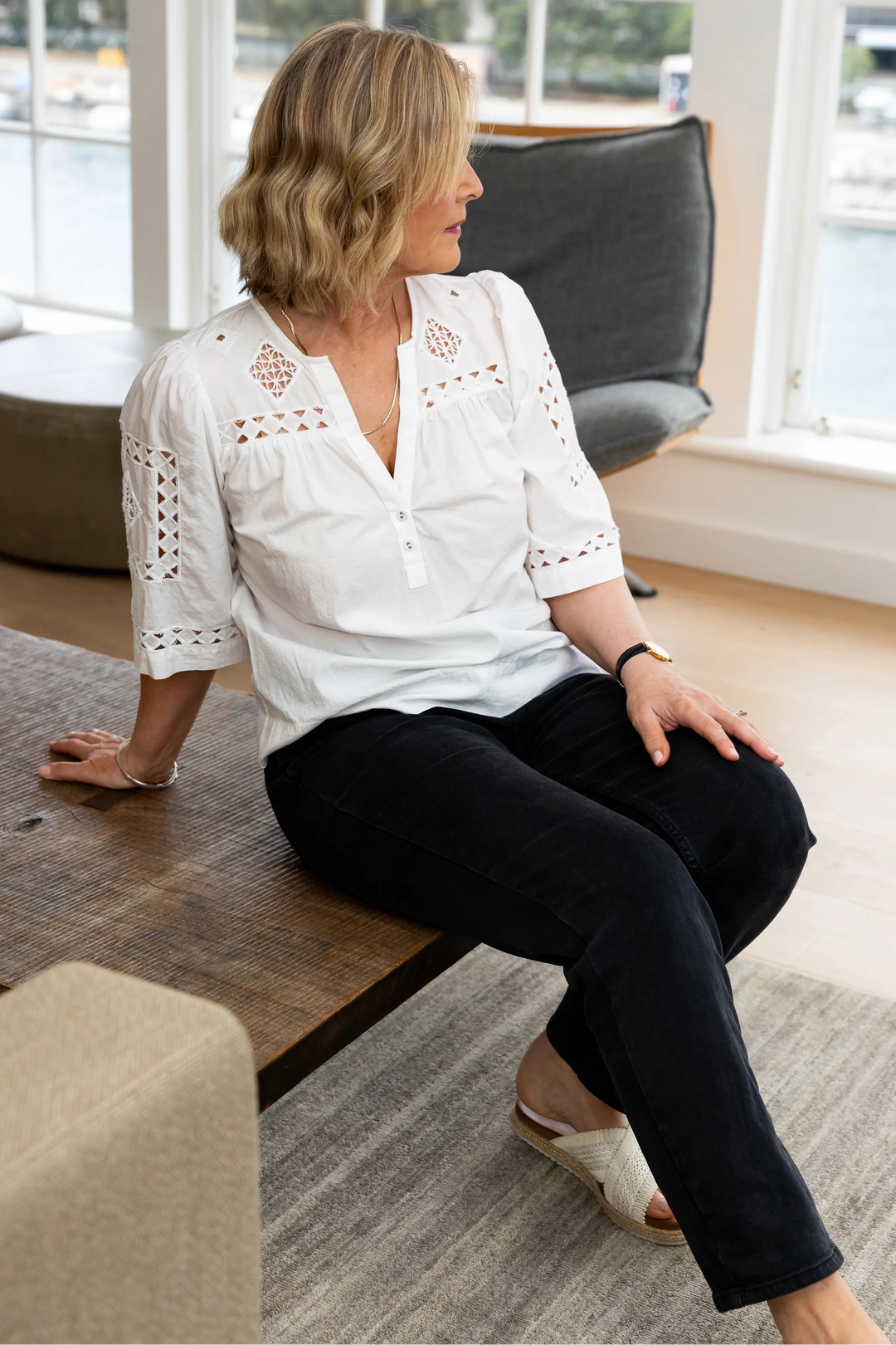 A woman with blonde hair sits on a wooden bench in a bright, modern room with large windows. She is wearing a white eyelet blouse with three-quarter sleeves and black pants.
