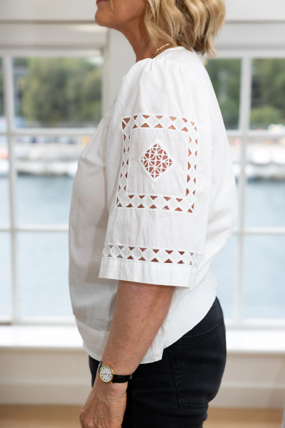 A woman wearing a white short-sleeve blouse with geometric cutout embroidery on the sleeve and hem, paired with black pants, standing in front of a bright window.