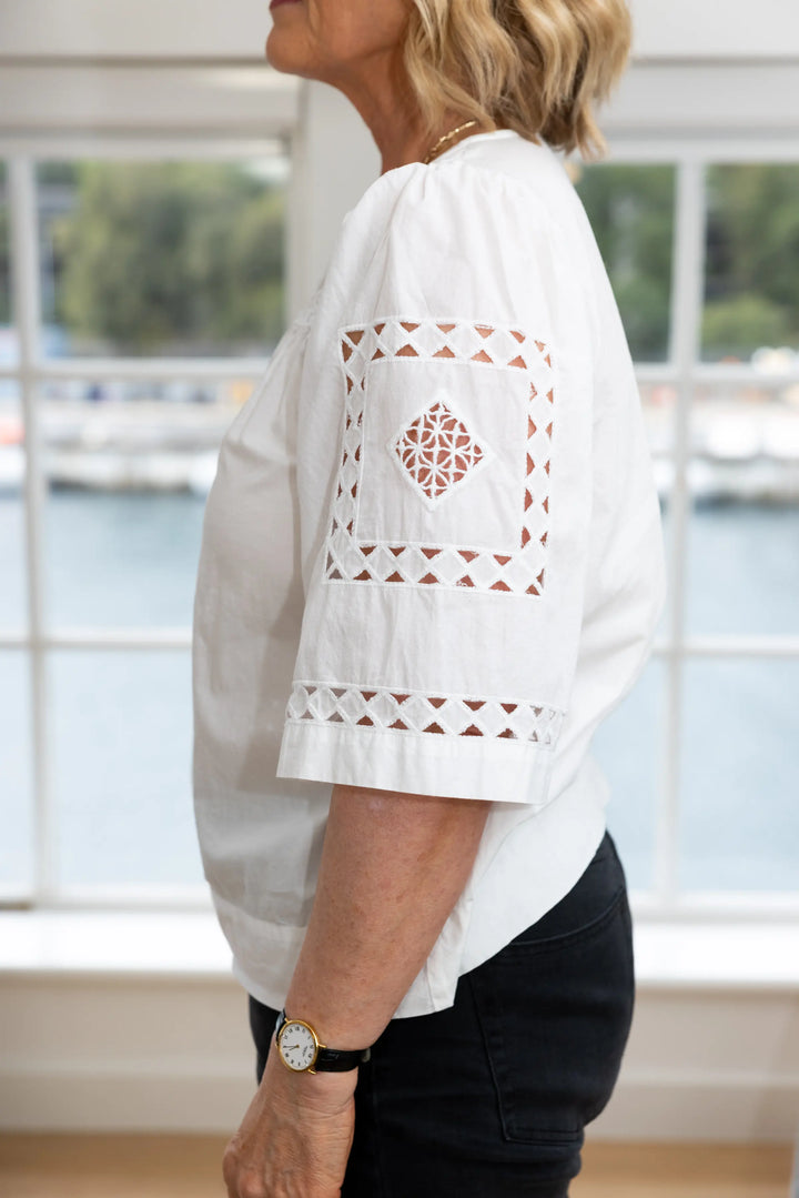 A woman wearing a white short-sleeve blouse with geometric cutout embroidery on the sleeve and hem, paired with black pants, standing in front of a bright window.