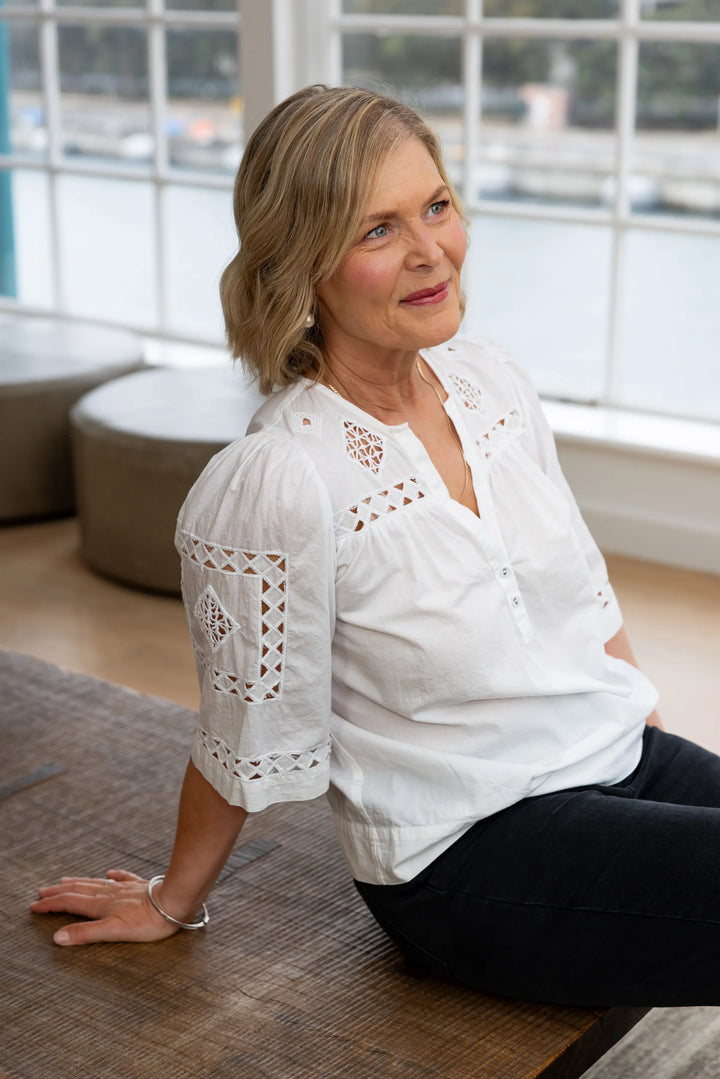 A woman with blonde hair sits on a wooden bench in a bright room, wearing a white short-sleeve blouse with intricate eyelet embroidery and black pants.