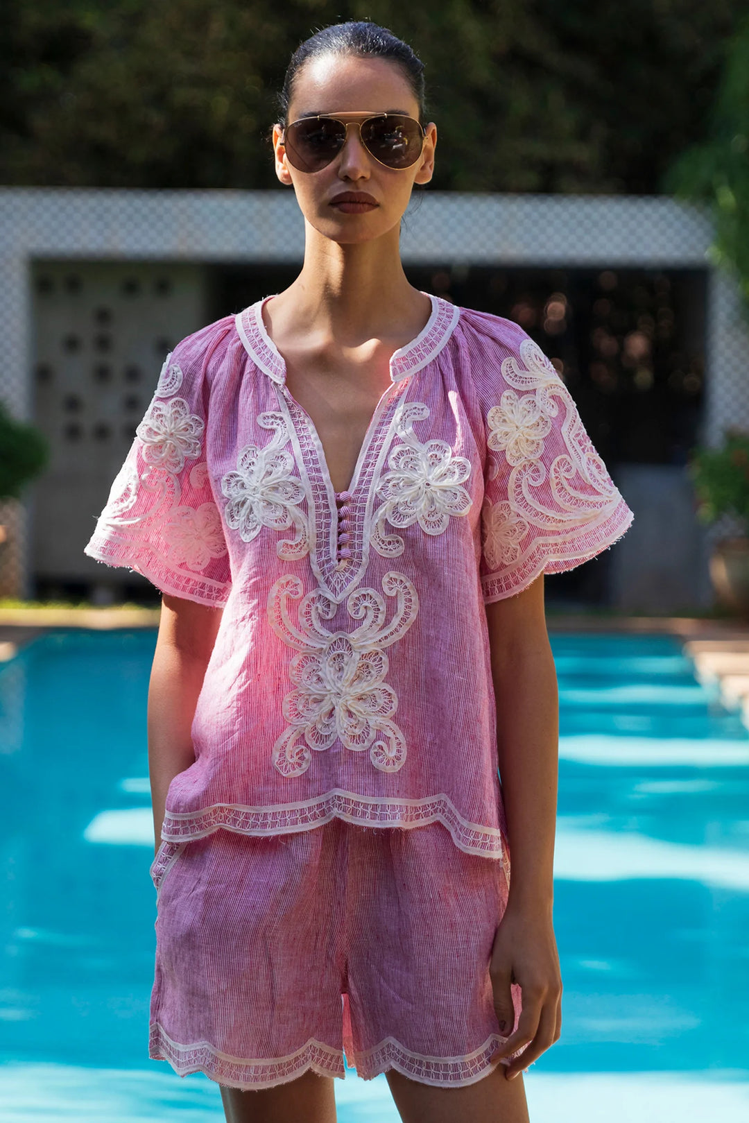 A woman wearing a pink embroidered lounge set stands in front of a swimming pool. The set includes a short-sleeve tunic top and matching shorts in a light pink fabric with white floral embroidery.