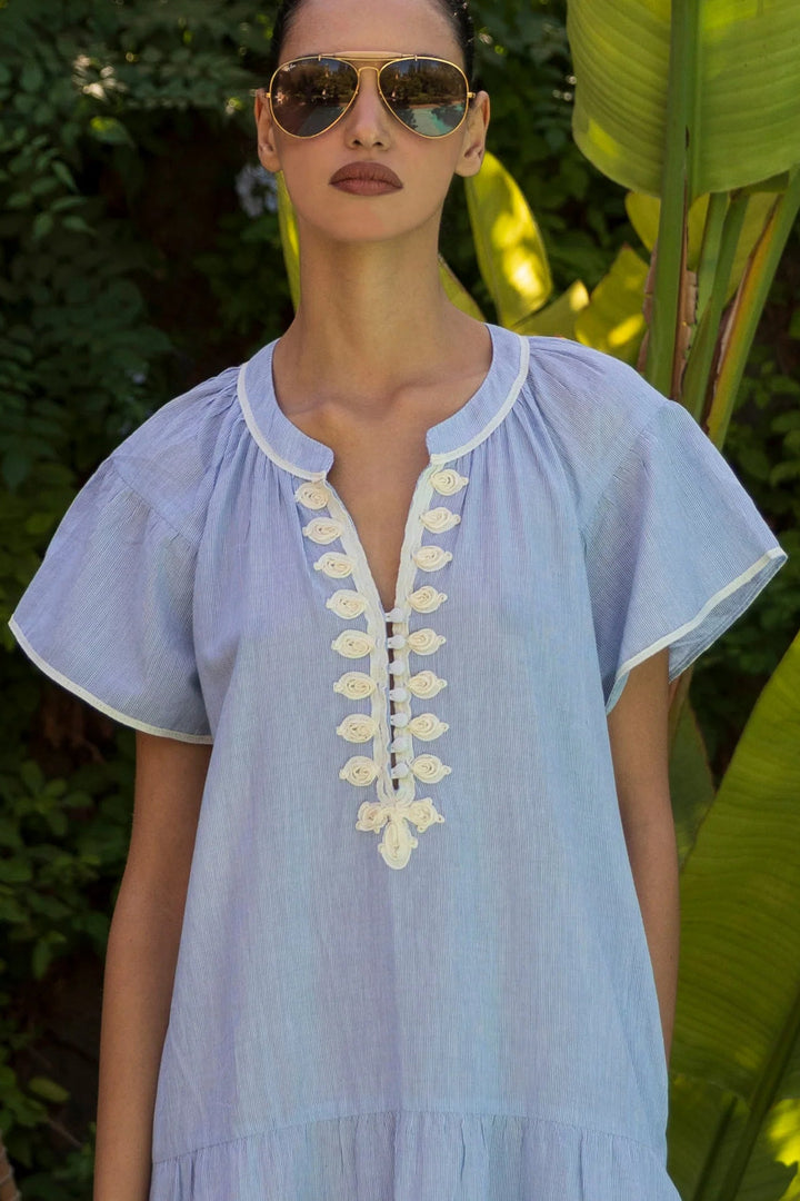 A woman wearing a light blue short-sleeve tunic dress with white trim and a decorative white floral lace-up front, photographed outdoors against a background of green tropical leaves.