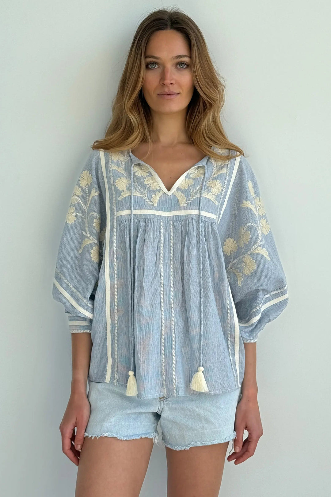 A woman modeling a light blue bohemian-style tunic top with cream embroidery, tassel ties, and three-quarter sleeves, paired with denim shorts against a plain light gray wall.