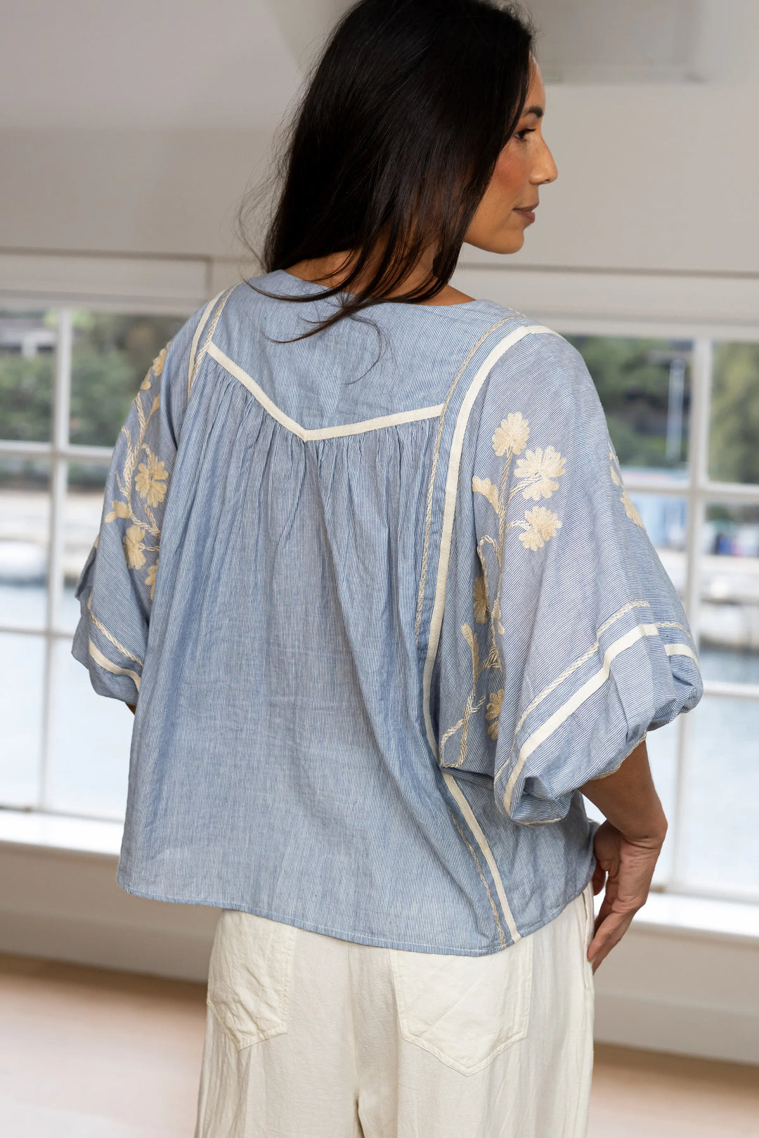 A woman shown from the back wearing a light blue chambray blouse with cream-colored piping and yellow floral embroidery on the sleeves, paired with cream-colored pants in a bright room.