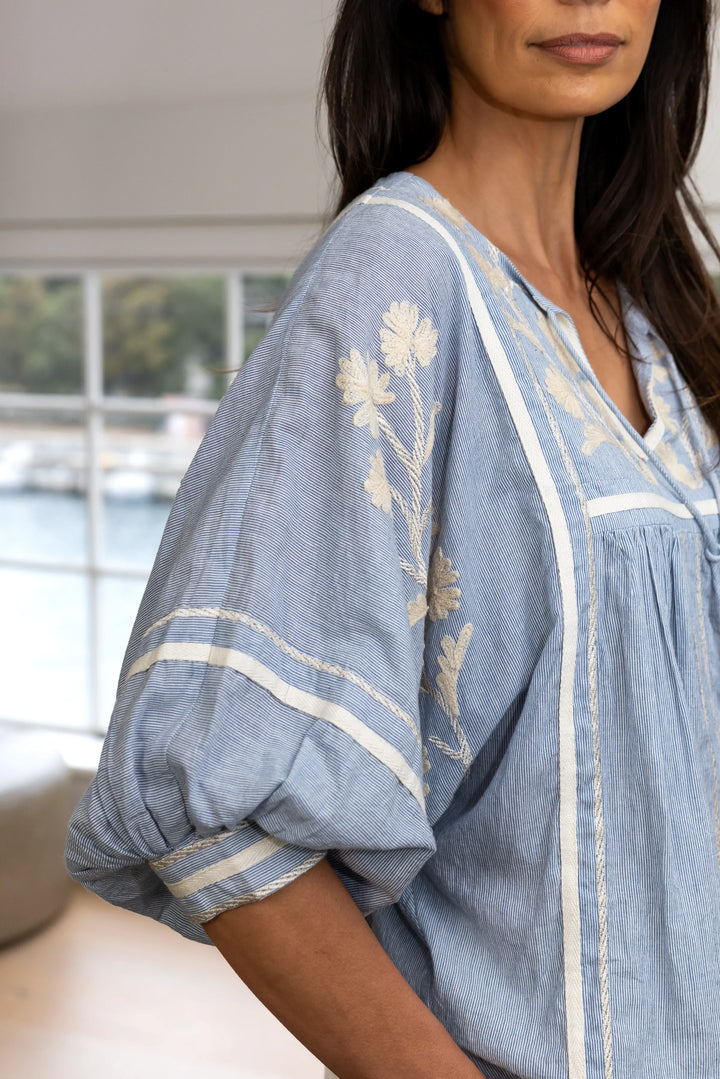 A close-up of a woman wearing a light blue cotton caftan blouse with cream-colored floral embroidery and striped trim, set against a softly blurred indoor background.