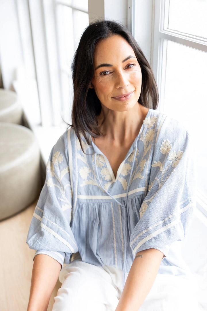 A woman with dark shoulder-length hair sits indoors near a bright window, wearing a light blue loose blouse with beige floral embroidery and white pants.