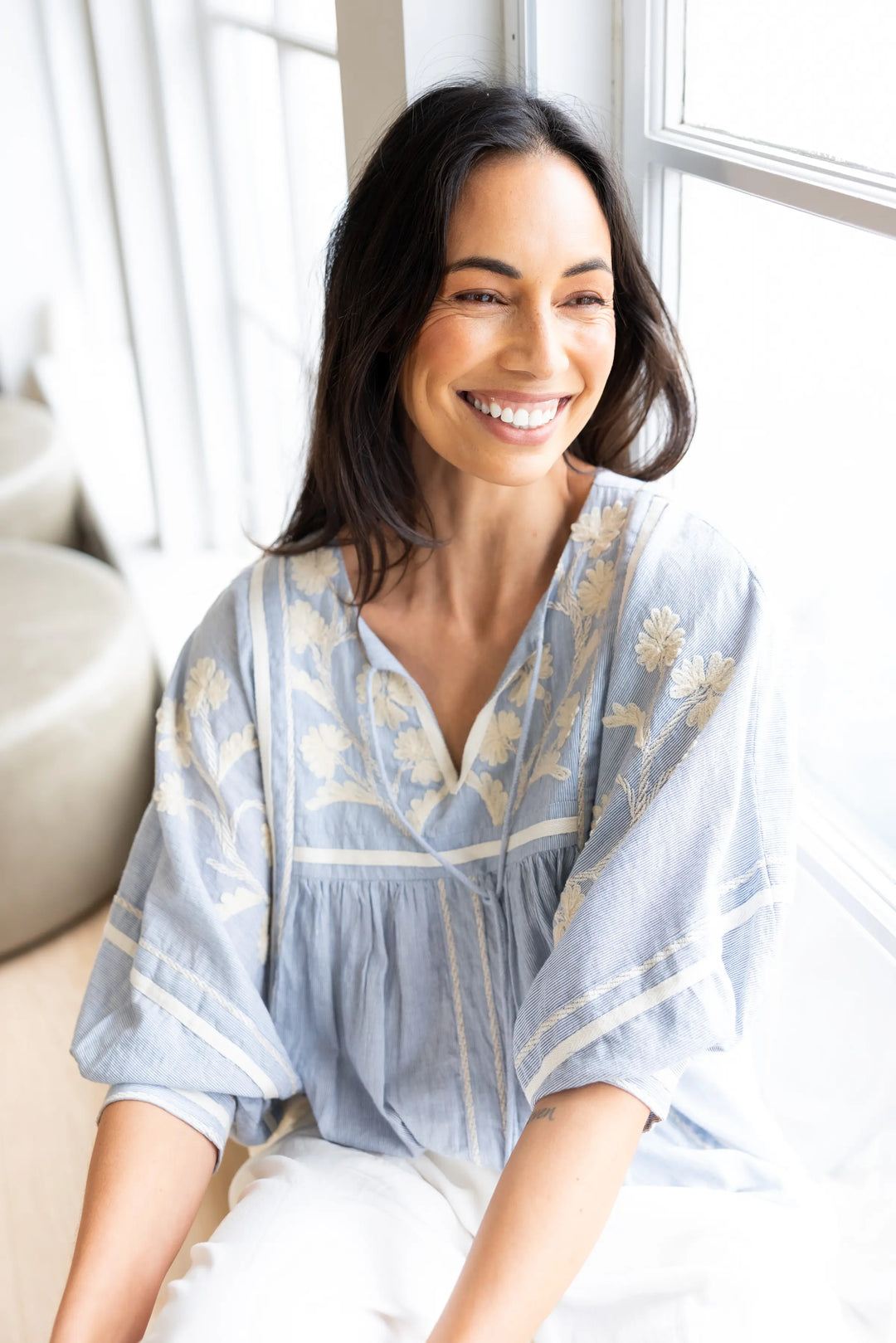 A smiling woman with dark hair sits indoors near a bright window, wearing a light blue bohemian-style blouse with beige floral embroidery and white pants.