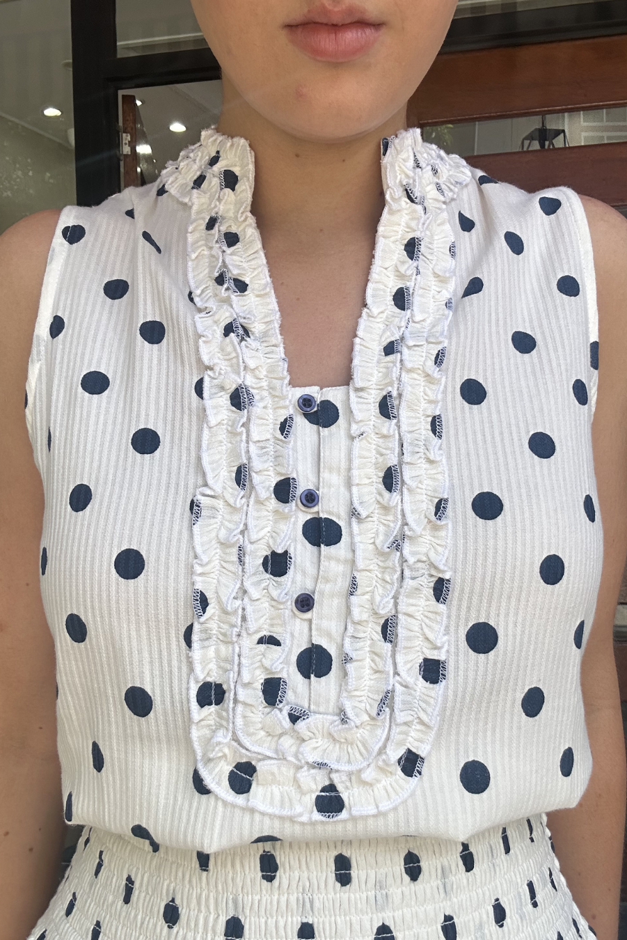 Person wearing a white sleeveless top with navy polka dots and ruffled neckline.