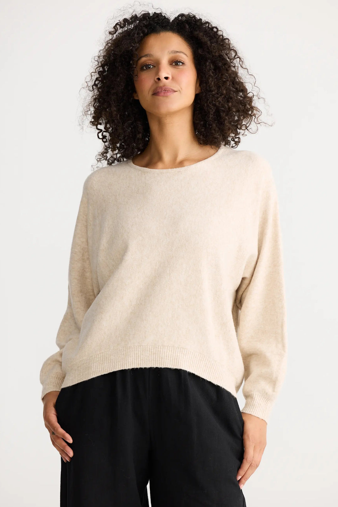 Woman wearing a beige sweater and black pants against a white background