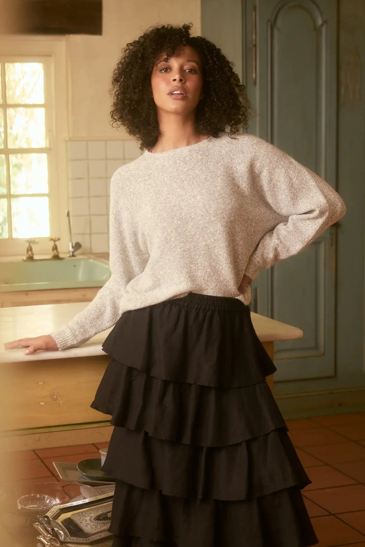 Woman standing in a kitchen wearing a white sweater and black tiered skirt.
