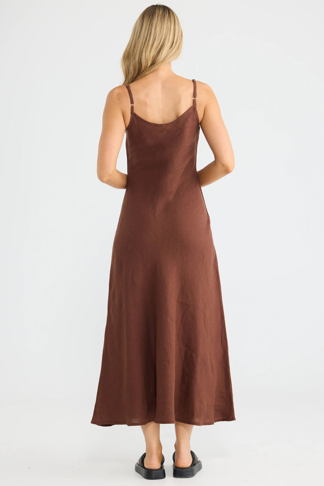 A woman viewed from the back wearing a brown spaghetti-strap midi slip dress, standing against a plain white studio background.