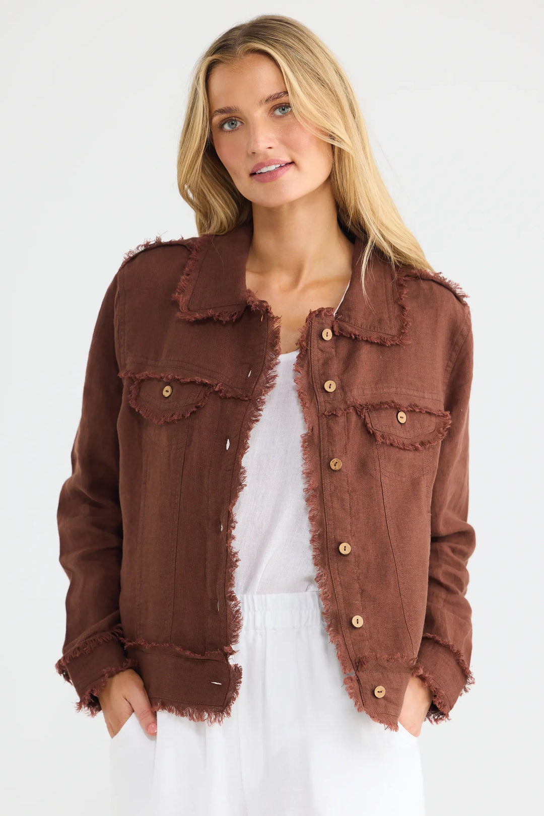 A woman modeling a brown frayed-edge denim jacket over a white top and white wide-leg pants, photographed against a clean white studio background.
