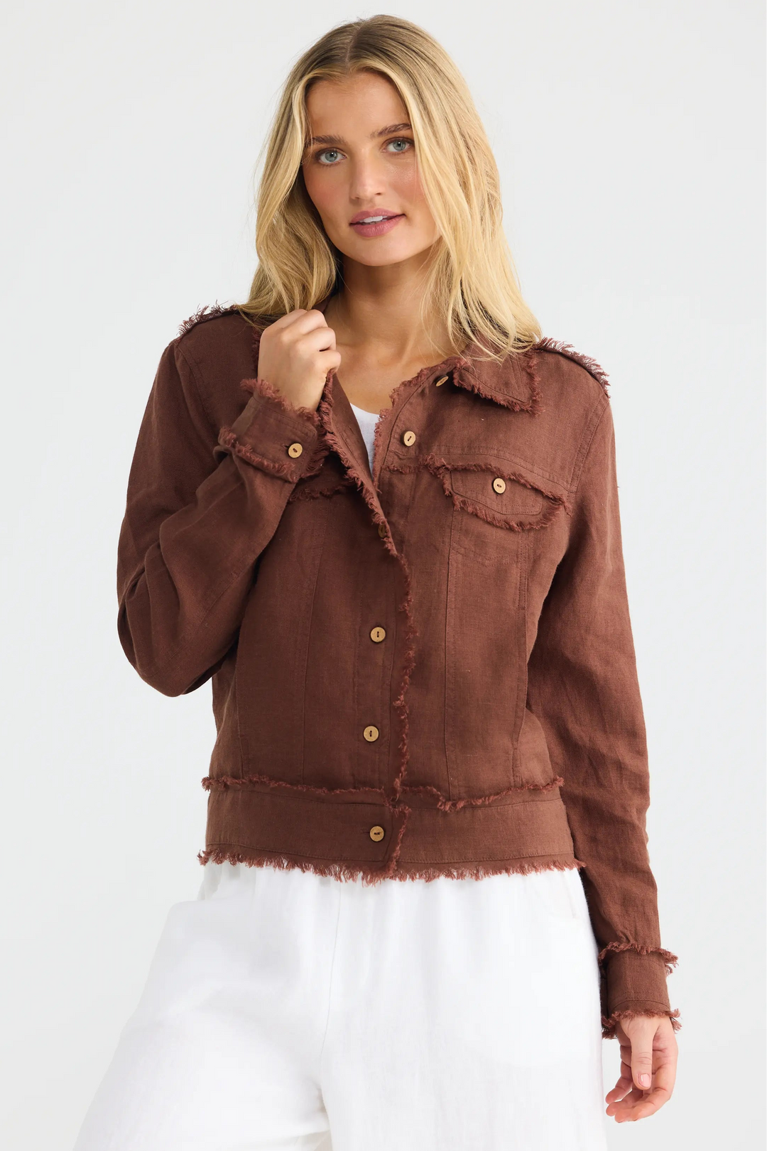 A woman modeling a cropped brown frayed-edge jacket with gold-tone buttons, paired with white pants, against a clean white studio background.