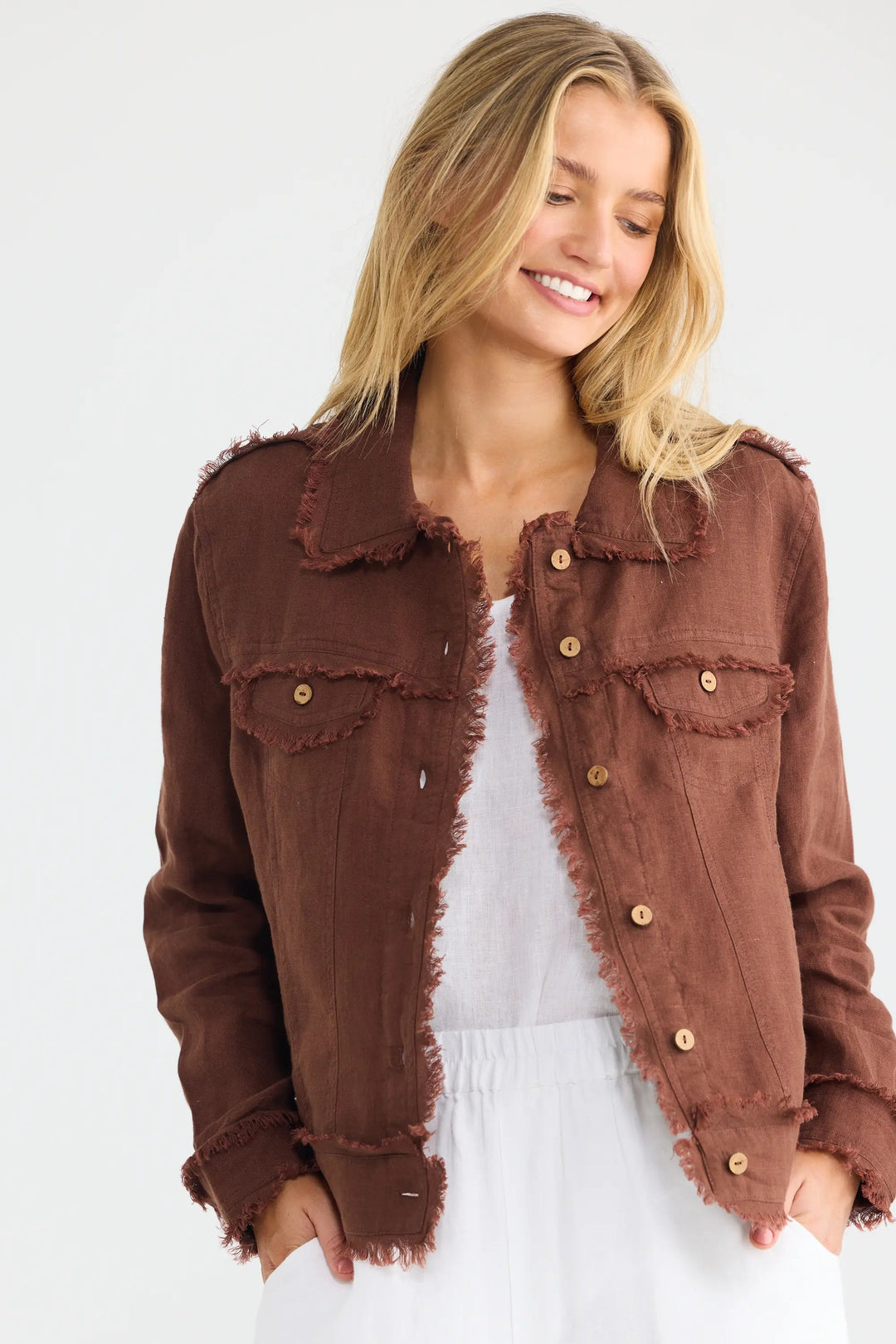 A woman wearing a brown frayed-edge denim jacket over a white top and white pants, standing against a plain white studio background.