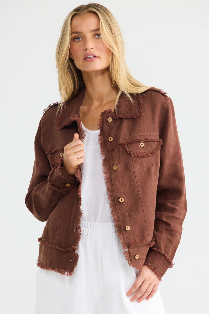 A blonde woman wearing a brown frayed-edge denim jacket over a white top and white wide-leg pants, standing against a plain white studio background.