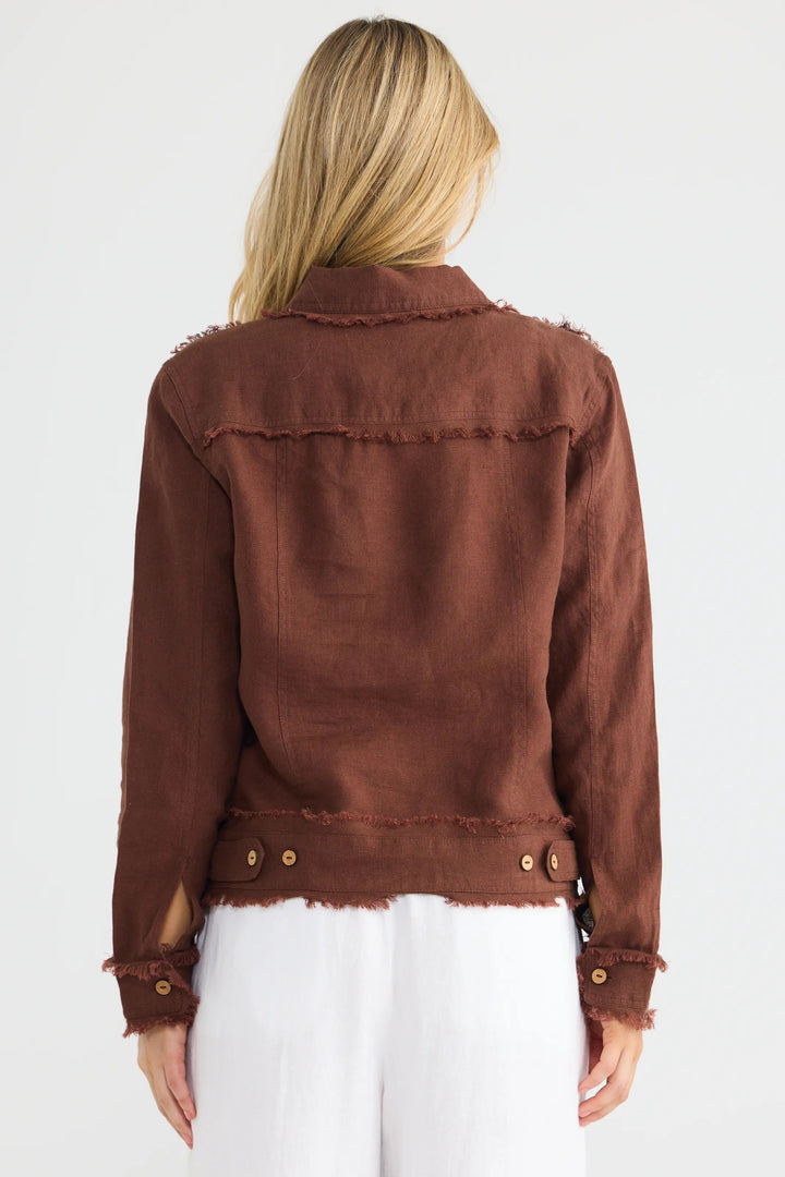 A woman viewed from the back wearing a brown frayed-edge jacket and white pants against a plain light gray studio background.