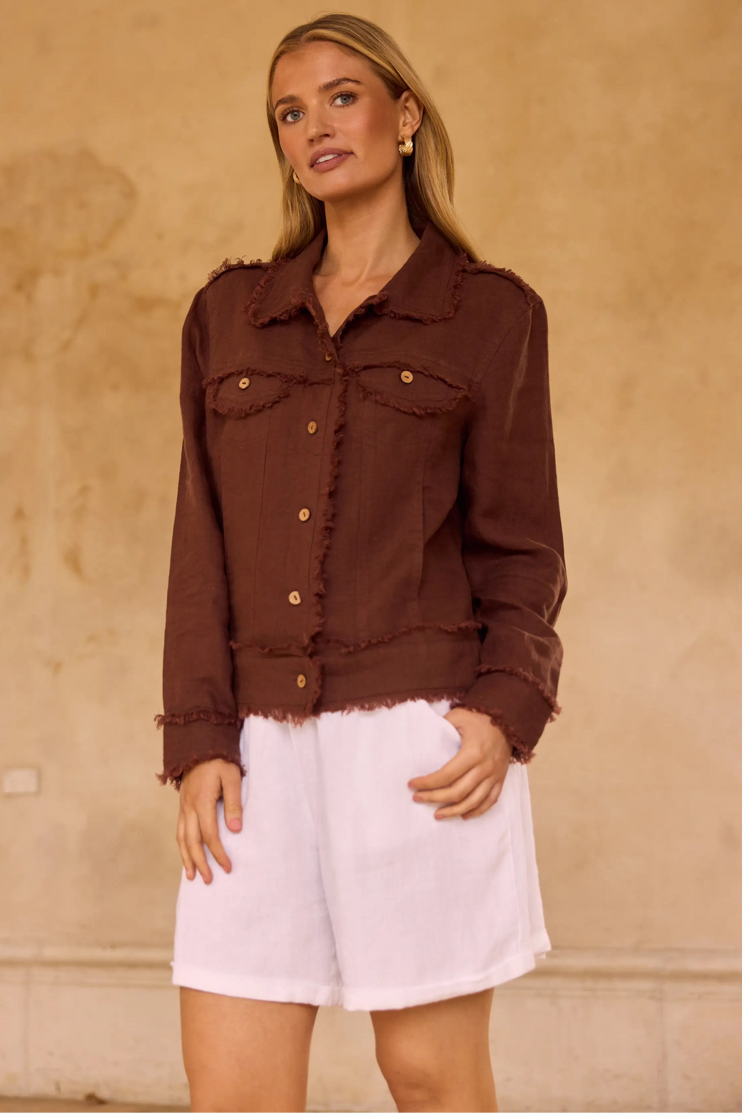 A woman modeling a dark brown frayed denim jacket with gold buttons, paired with white shorts, standing against a textured beige wall.