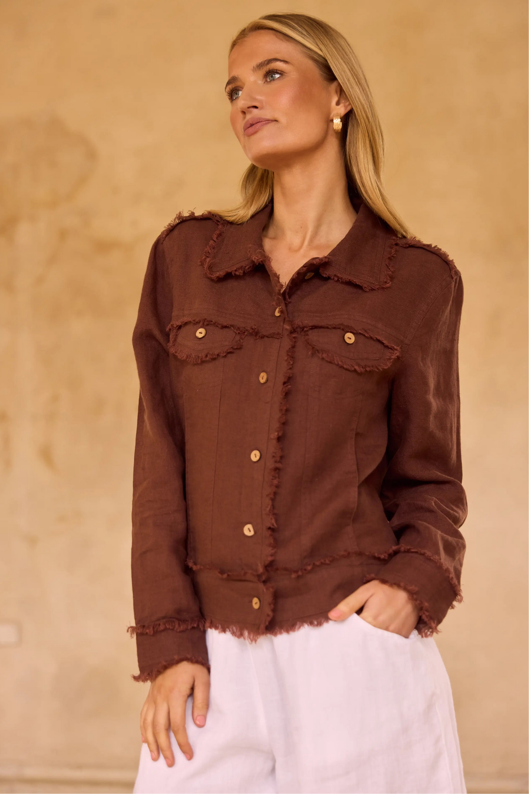 A woman wearing a brown frayed-edge button-up shirt jacket with gold-tone buttons, paired with white pants, standing against a textured beige wall.