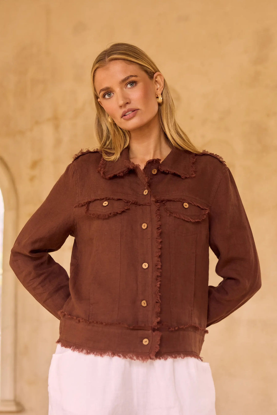 A woman modeling a brown frayed denim jacket with gold buttons, standing in a warm beige interior with a rustic wall and an arched window in the background.