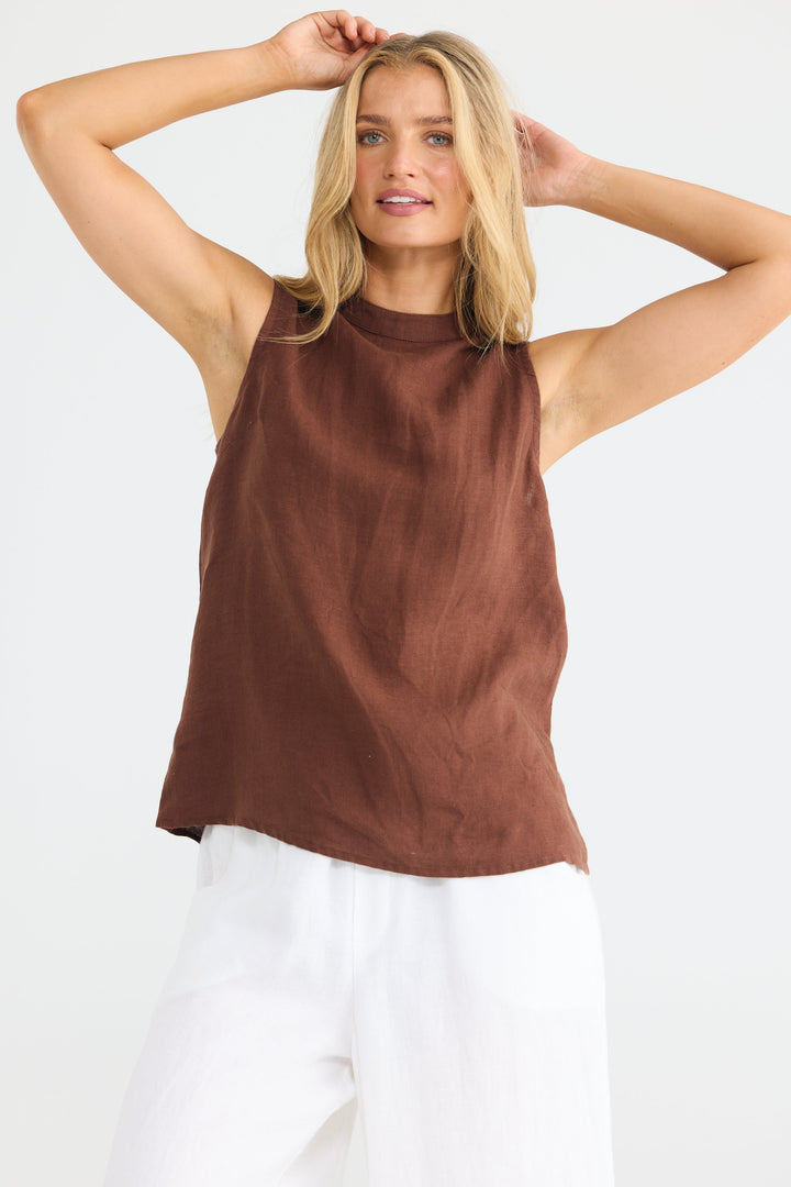 A woman modeling a sleeveless brown top with a high mock neck, paired with white pants, against a plain white studio background.