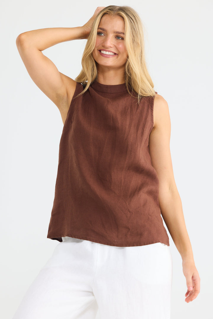 A woman modeling a sleeveless brown top with a mock neck and white pants against a plain white studio background.