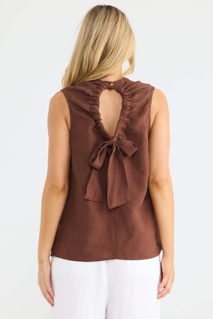A woman viewed from the back wearing a sleeveless brown top with a large bow detail at the back neckline and white pants, against a plain light background.