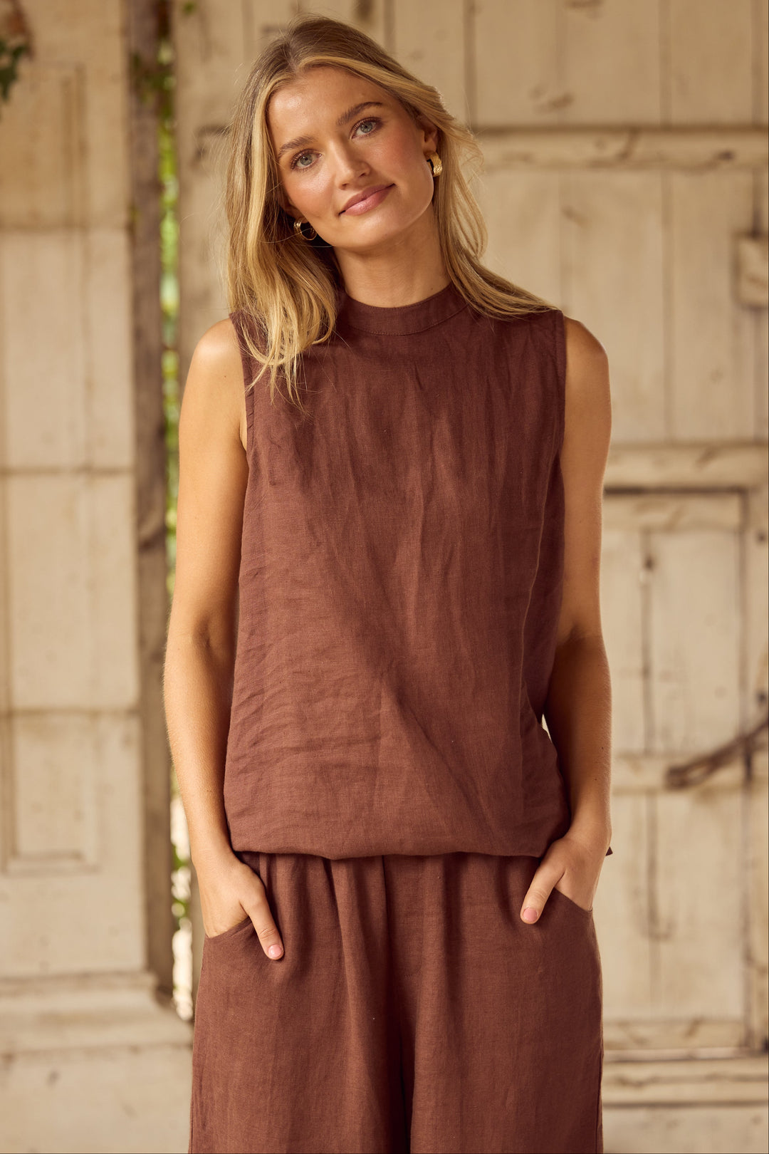 A woman wearing a sleeveless rust-brown linen top and matching wide-leg linen pants, standing in front of a rustic light-colored wooden door.